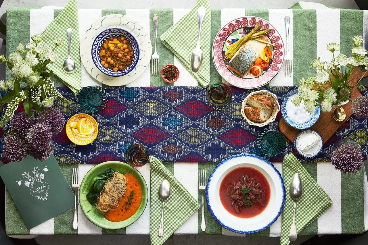 A table of organic, local, gluten free New York City flexitarian, pescatarian, carnitarian, vegan, and vegetarian plated meal delivery foods that has two plates of soup, a bowl of gluten free bread, two spoons, purple flowers, green napkins, and green menu on a table